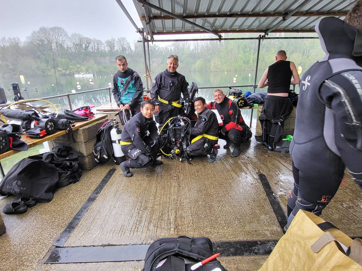 Instructor candidates in drysuits kitting up under the shelter on a quarry pontoon, smiling at the camera