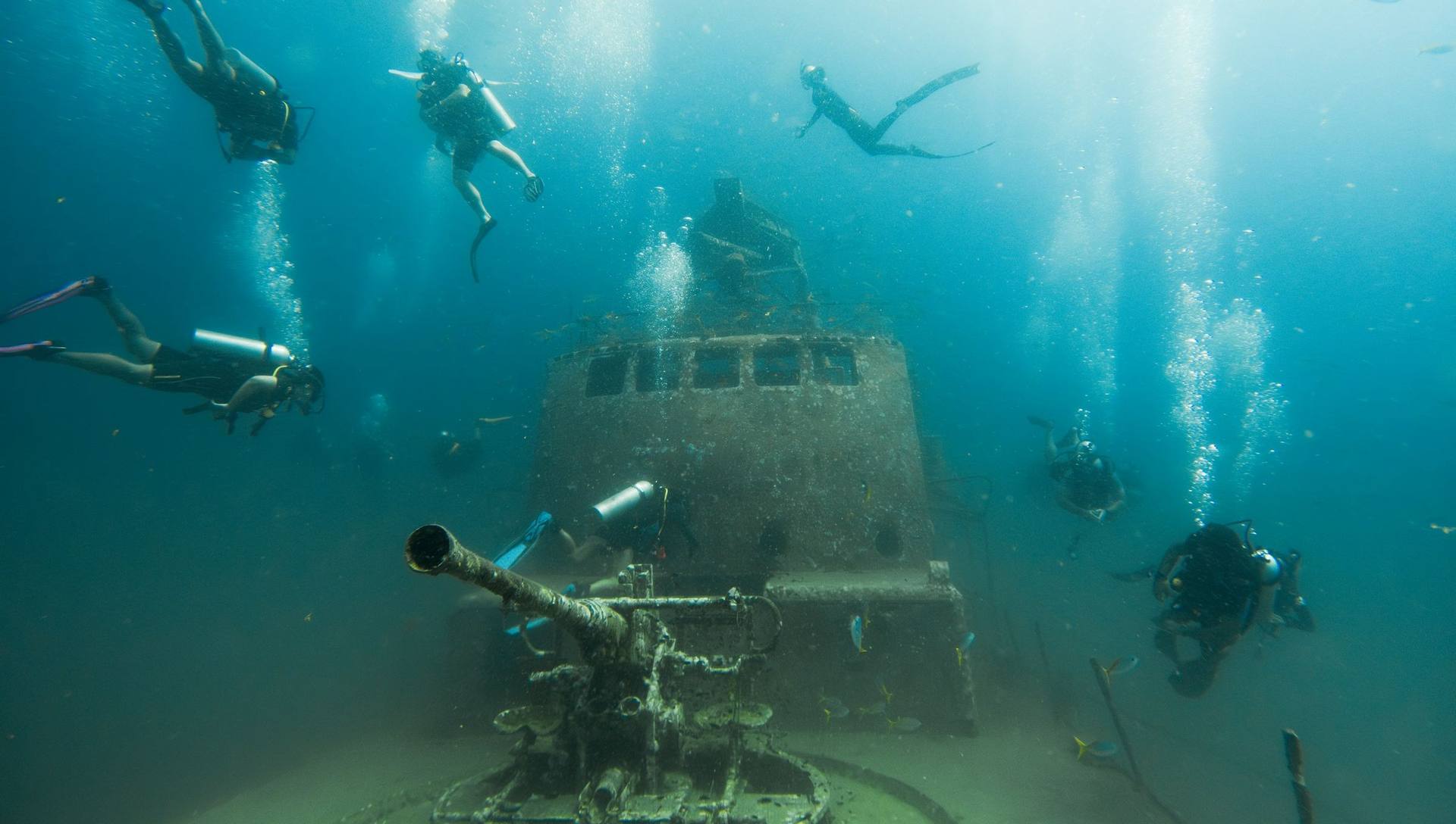Scuba divers exploring a sunken patrol boat with a deck gun, bubbles rising towards the surface