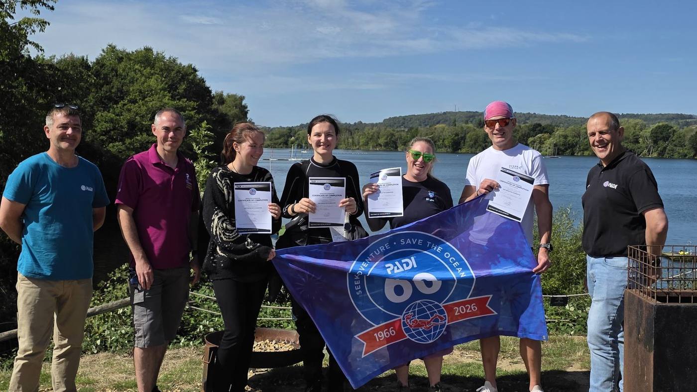 A group of new instructors holding certificates and a PADI 60th anniversary flag by the lake