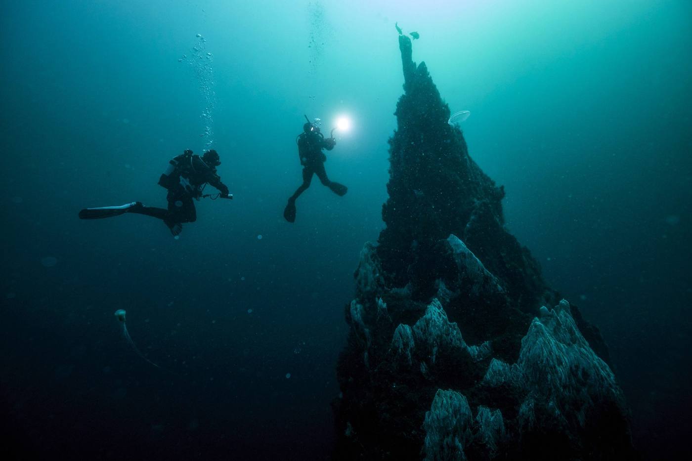 Two divers with a torch descending beside a deep rock pinnacle in blue water