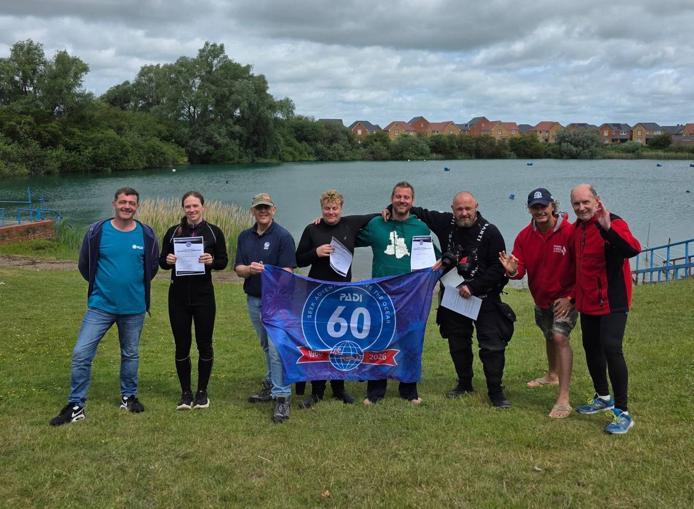 Instructor candidates and staff by a lake holding certificates and a PADI 60th anniversary flag