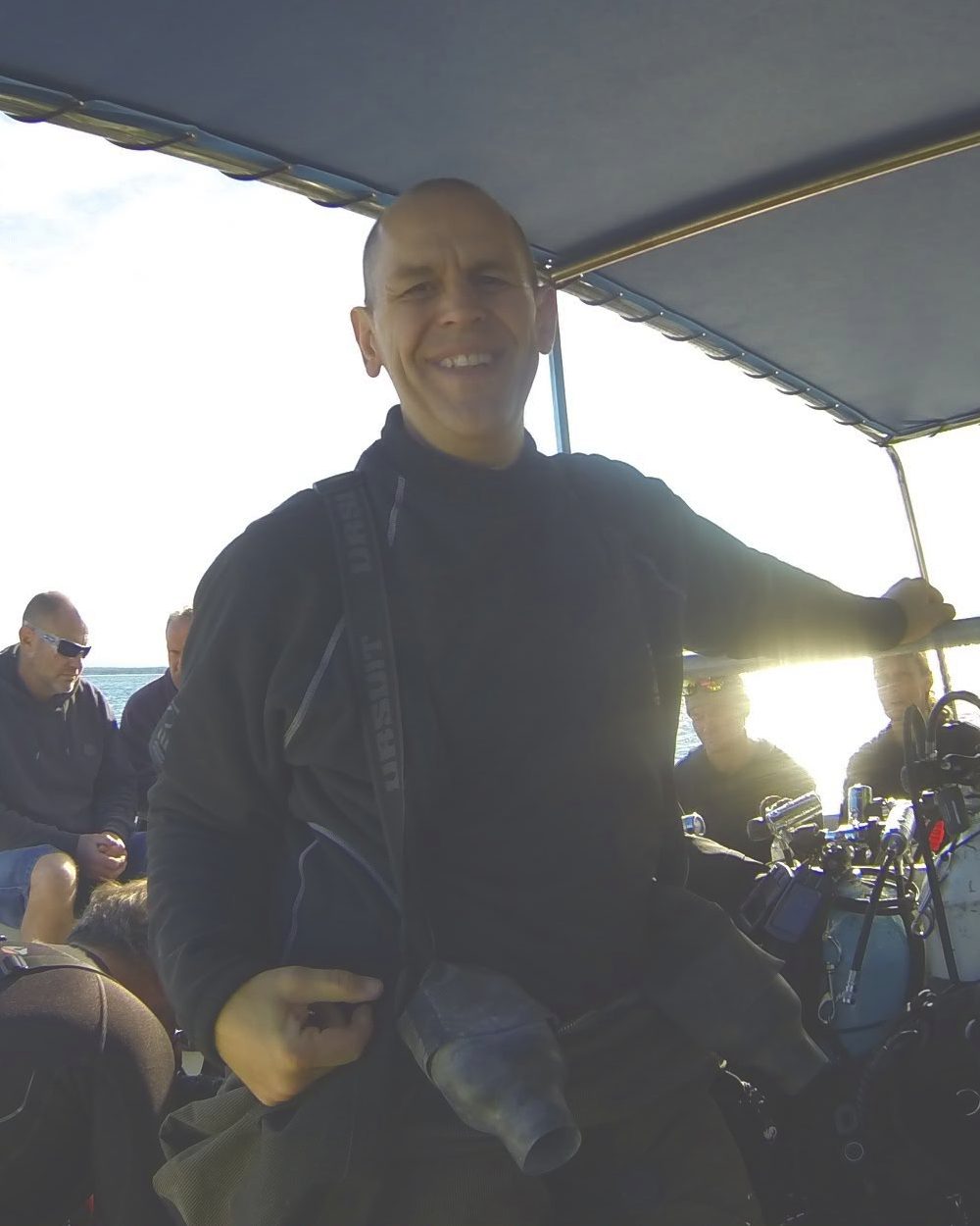 Scott Roberts smiling on a dive boat in his drysuit undersuit, with divers and kit behind him