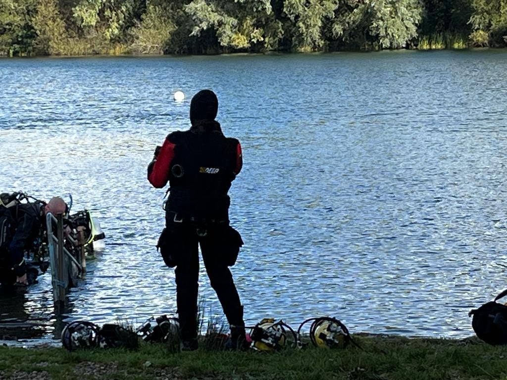 A drysuit diver standing at the water's edge with regulators laid out on the grass